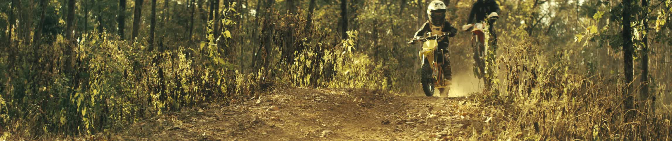 Two man wearing full protective gear and a helmet rides a yellow dirt bike down a dusty trail through a dense forest as sunlight filters through the trees