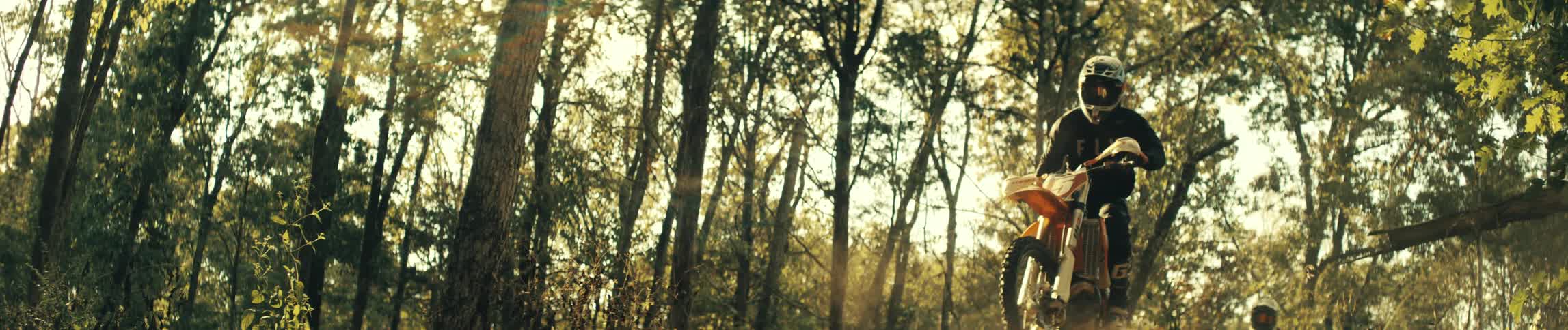 A person wearing a black jersey and a helmet rides an orange dirt bike through a wooded area with tall trees as golden sunlight filters through the branches and dust rises from the ground