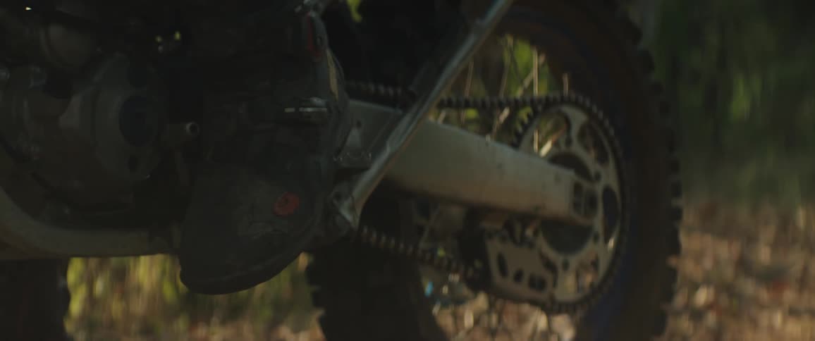 A person rests their black riding boot on a foot peg while the camera focuses on the dirty chain and rear sprocket of a stationary dirt bike parked on a wooded trail during the golden hour