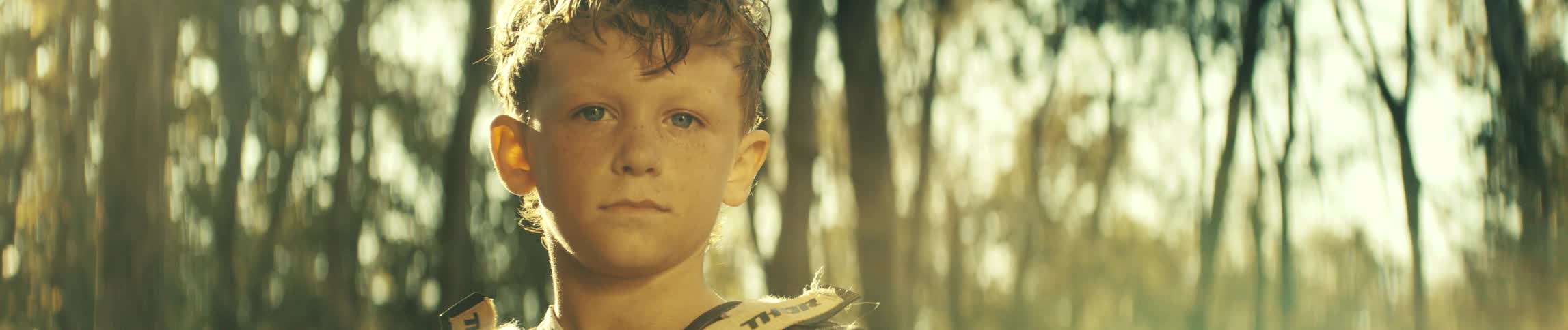 Boy looks into the camera as he stands in the woods