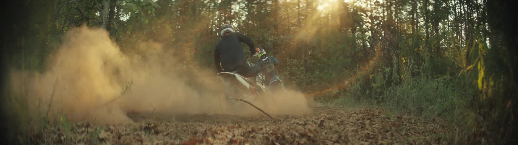 Two motocross riders lean into a turn on a dirt trail in a forest kicking up dust as sunlight filters through the trees