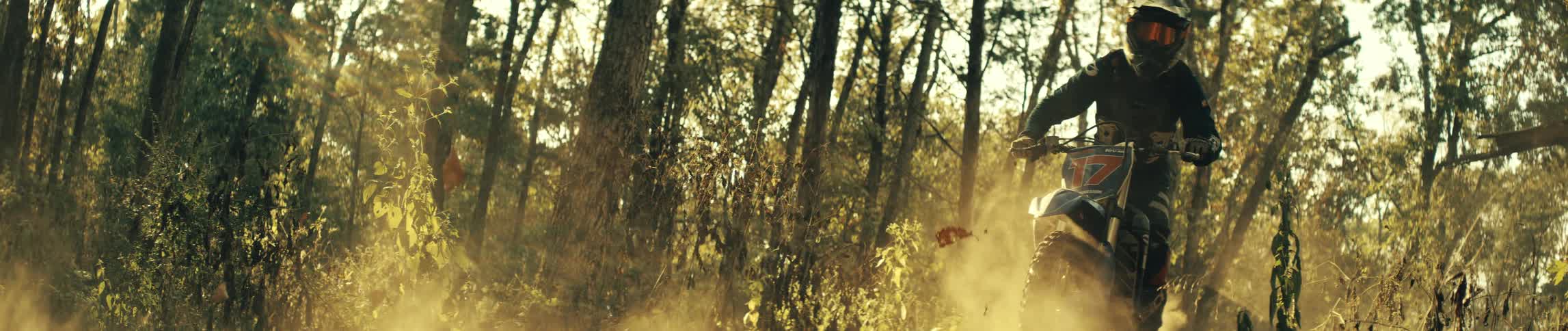 Two riders ride dirt bikes on a forest trail under sunlight with dust rising along the path
