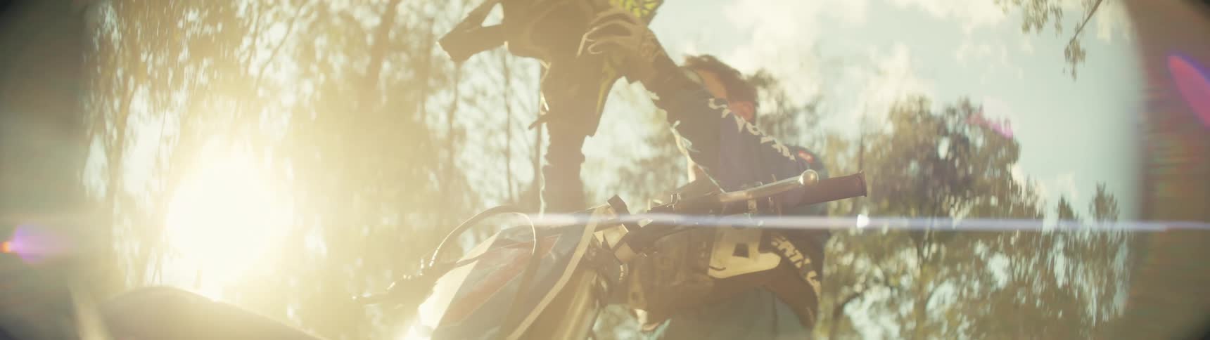 A man in blue riding gear stands behind his motocross bike and pulls a black helmet over his head while bright sunlight creates a warm flare across the outdoor wooded setting