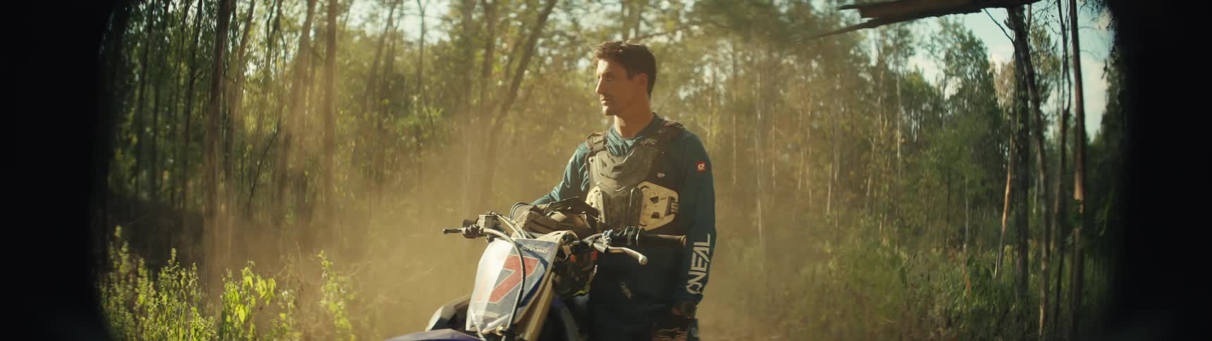 A mountain biker in the woods stands beside his bike wearing a blue jacket at midday