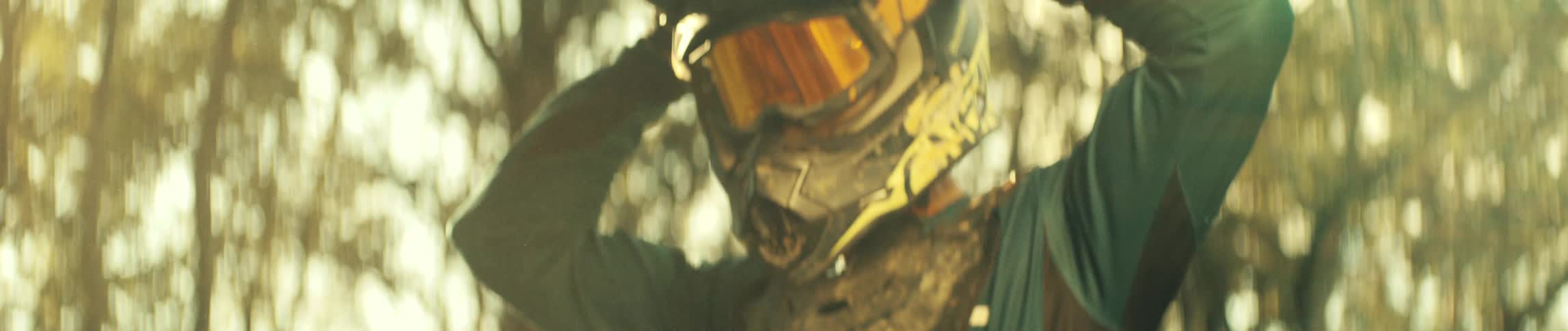 A closeup shows a motocross rider wearing a helmet and places on his face reflective goggles in warm golden forest light