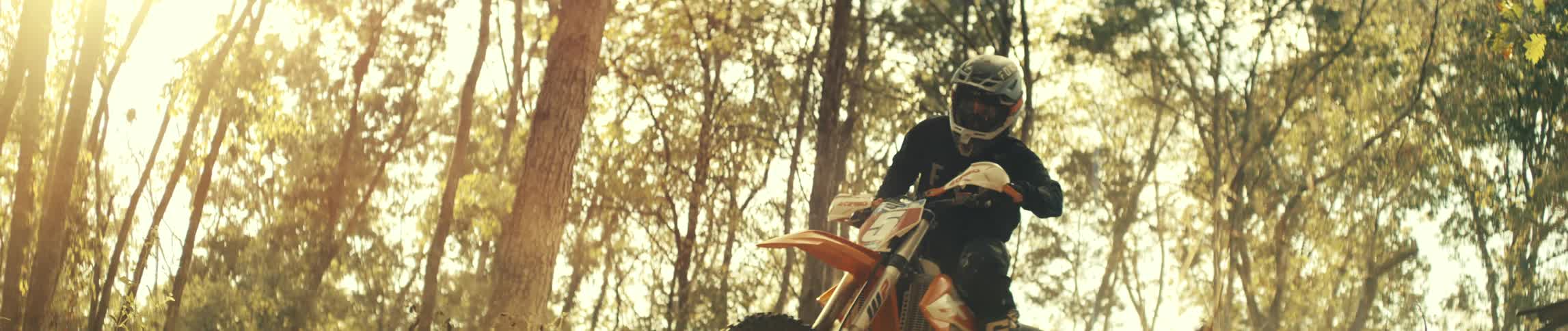 A motocross rider launches over a small rise in a sunlit forest the orange bike lifting into the warm golden air