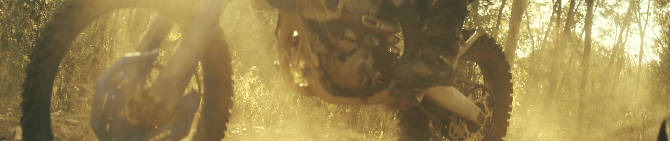 Front wheel of a dirt bike moves along a forest trail under strong sunlight and suspended dust