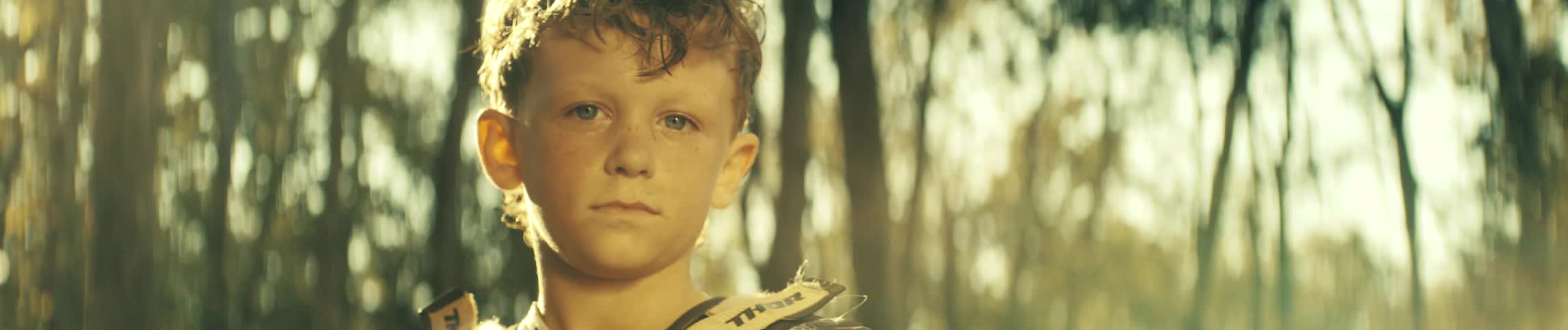 A young boy with freckles and curly hair stares directly into the camera with a serious expression while wearing motocross gear in a sunlit forest