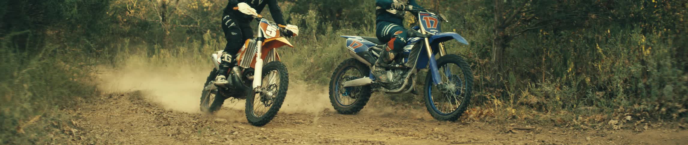 Two people drive dirt bikes along a dirt trail at a high speed