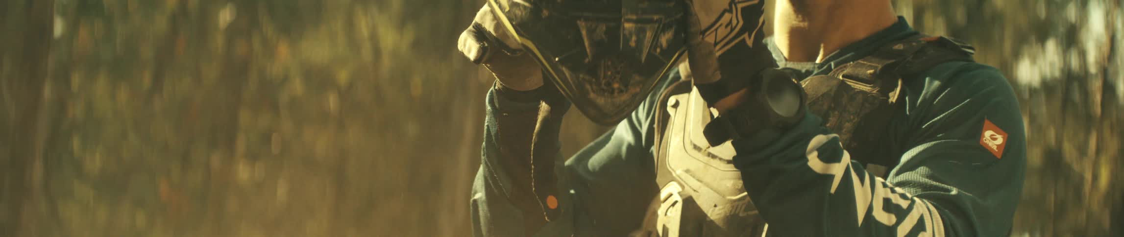 A motocross rider wearing gloves and chest armor slowly pulls a black mudsplattered helmet over their headthe scene is shot in a warm dusty environment with golden sunlight filtering through the background
