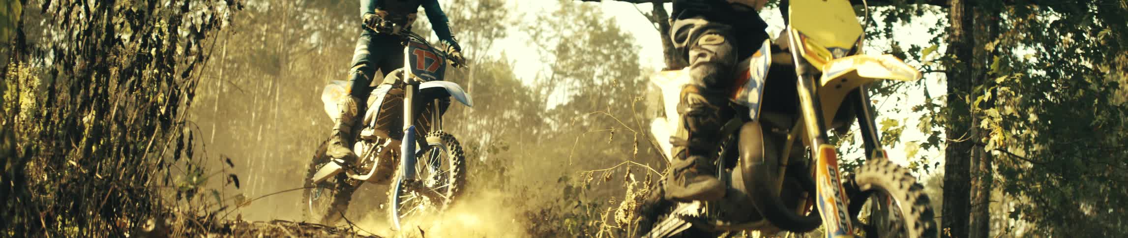 Two riders ride dirt bikes on a forest trail under sunlight one of them performing a jump