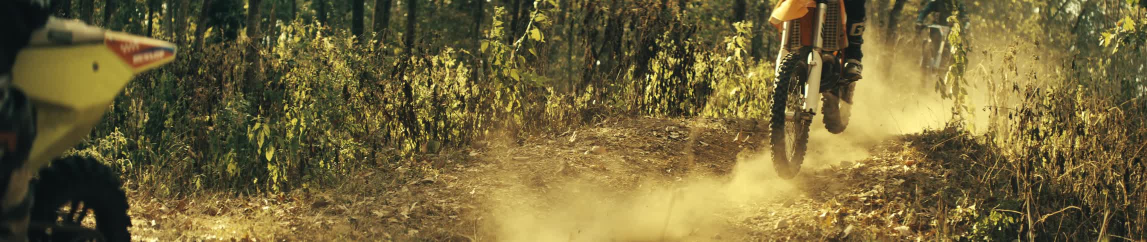 Three riders wearing protective gear and a helmet rides dirt bikes through a sunny forest trail kicking up a cloud of dust from the ground while navigating a slight turn among the trees and dense undergrowth