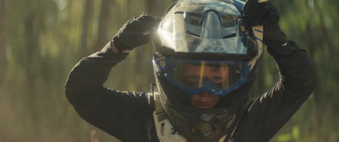 A young boy wearing full motocross gear sits on a yellow dirt bike in a wooded area and pulls his goggles down over his eyes while looking directly into the camera