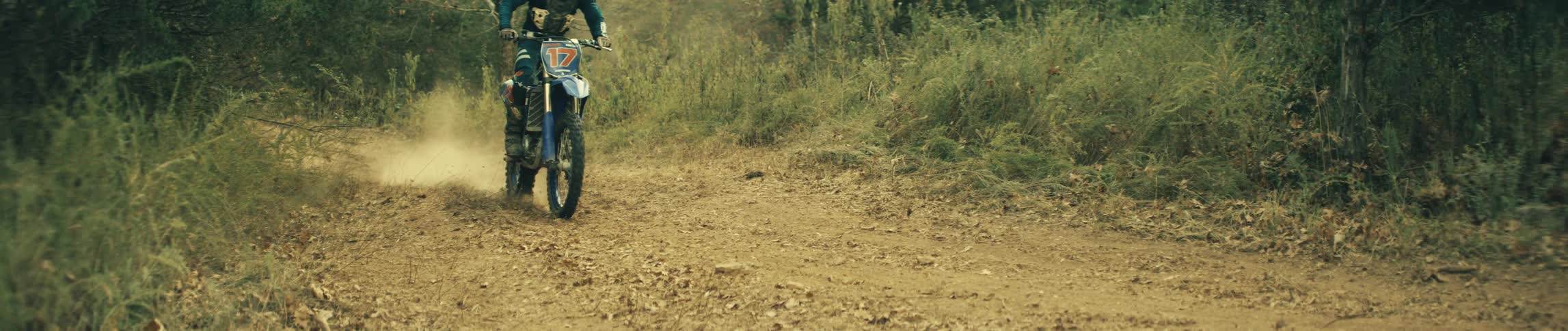Person rides a dirt bike on a forest dirt trail kicking up dust behind the rear wheel