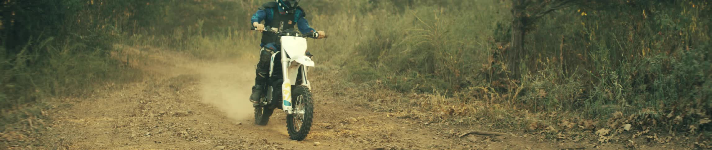 A young rider in blue gear and a helmet navigates a white dirt bike down a dusty unpaved paththe camera tracks low as the bike kicks up dirt eventually focusing on the front wheel as it lifts slightly off the ground
