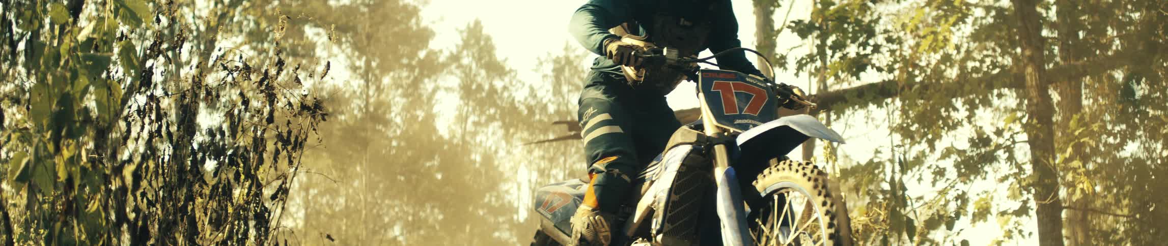 A motorcyclist wearing blue gear and a helmet jumps a blue dirt bike through the air over a wooded trail while dust kicks up from the ground below in the hazy golden sunlight