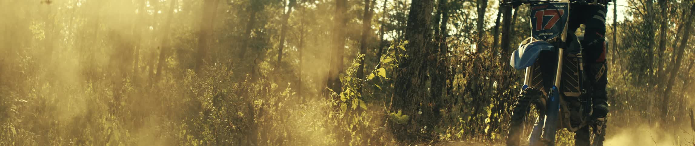 Person rides a dirt bike on a forest dirt trail under sunlight with dust in the air