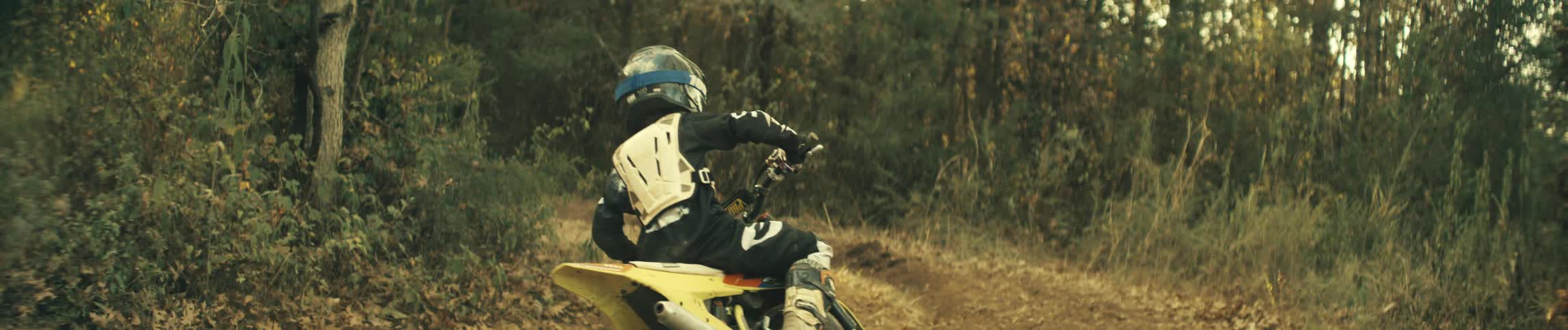 A young rider wearing protective gear accelerates a yellow dirt bike on a rural trail kicking up a large cloud of brown dust from the dry ground