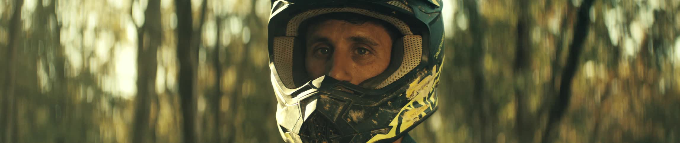 Man wears a motocross helmet in an outdoor setting with trees in the background