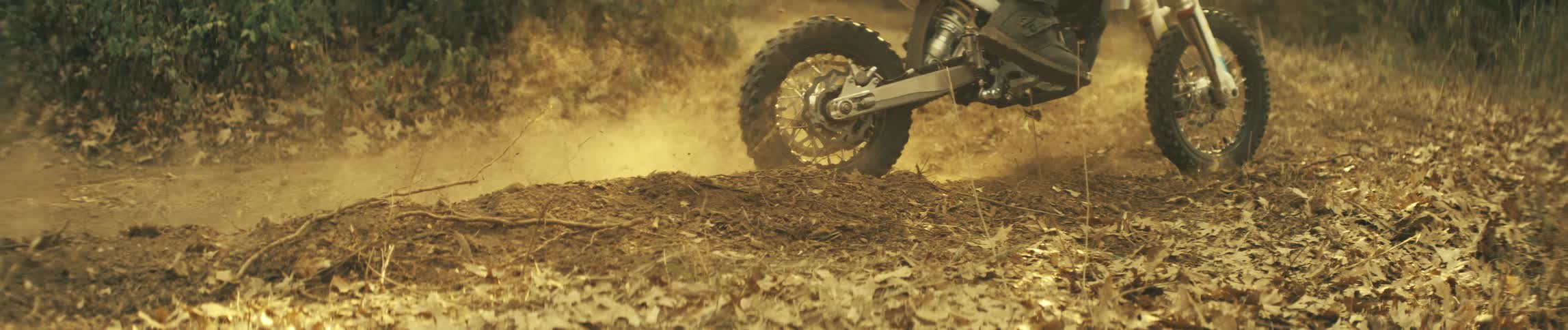 A dirt bikes front wheel skims over a small mound of earth scattering dry leaves across the forest floor
