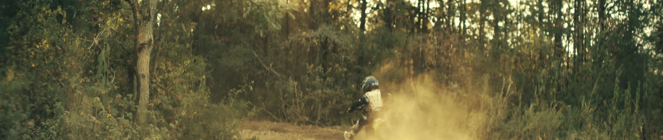 A man wearing a blue helmet and white chest protector rides a yellow dirt bike down a dusty trail through a wooded area while kicking up a thick cloud of brown sand behind the rear tire