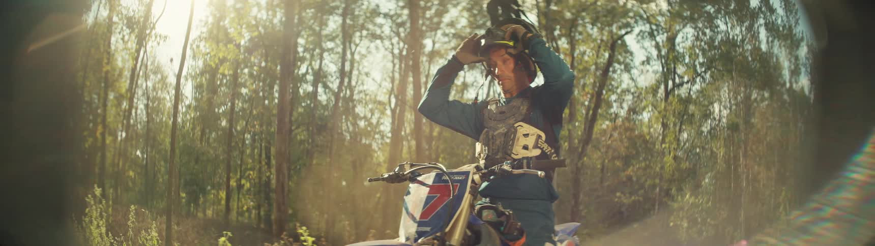 A man takes off his helmet while seated on a dirt bike in a forest