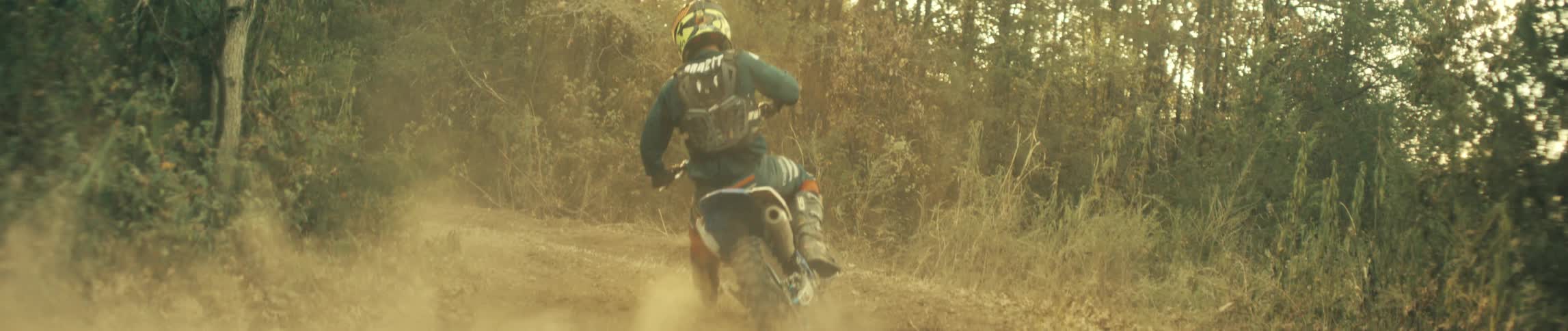 Person rides a dirt bike around a bend on a forest dirt trail kicking up dust