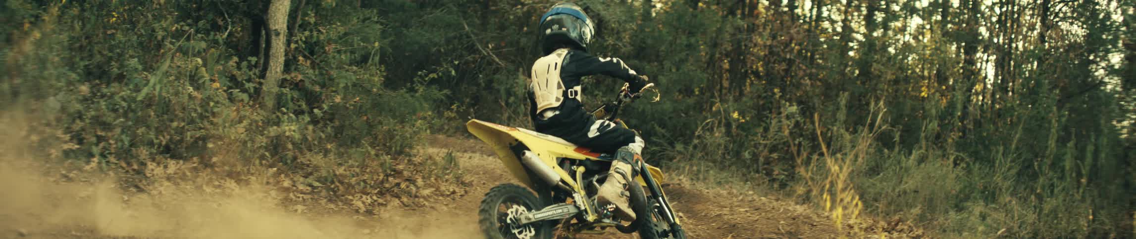 A motocross rider wearing a white chest protector and blue helmet accelerates a yellow dirt bike down a dusty trail toward a wooded area