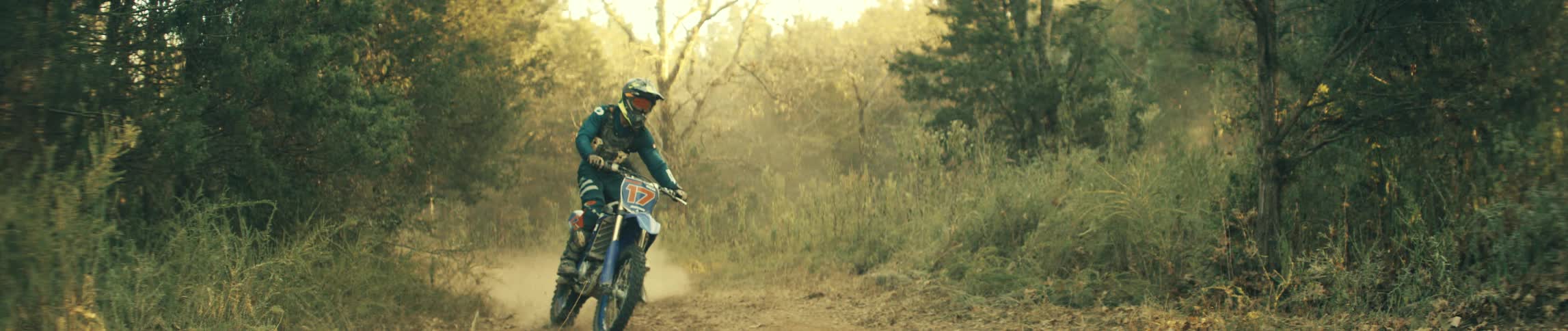 Two people ride dirt bikes through a sunlit forest kicking up dust as golden light filters through the trees