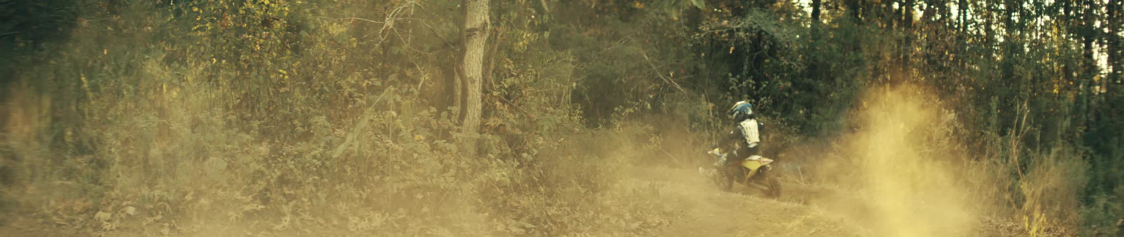 A young motocross rider speeds away from view on a yellow dirt bike down a dusty trail kicking up a thick golden cloud of dirt before disappearing into the sunlit forest
