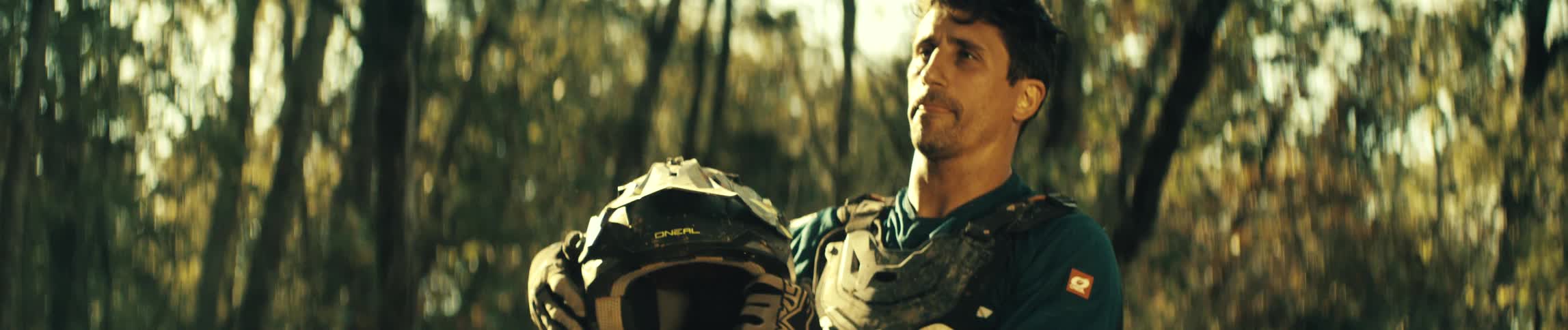 Man removes his motocross helmet in the middle of a sunlit forest