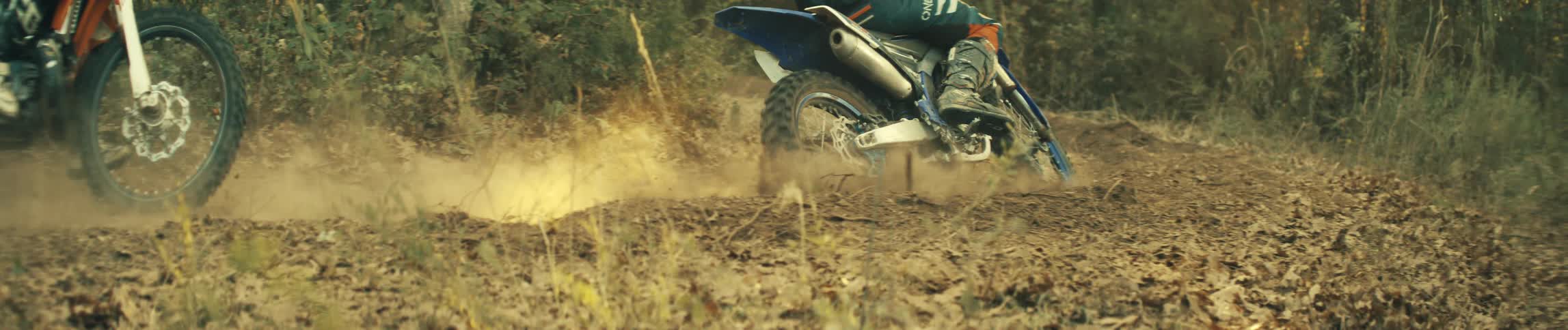 Two riders drive dirt bikes on a dirt trail kicking up dust