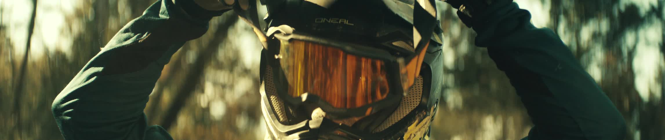 A person is in the woods seen from the chest up putting on googles to go over their motor cross helmet