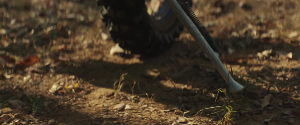 A closeup shows a dirt bikes metal footpeg and engine as a riders boot rests firmly in place over the forest floor