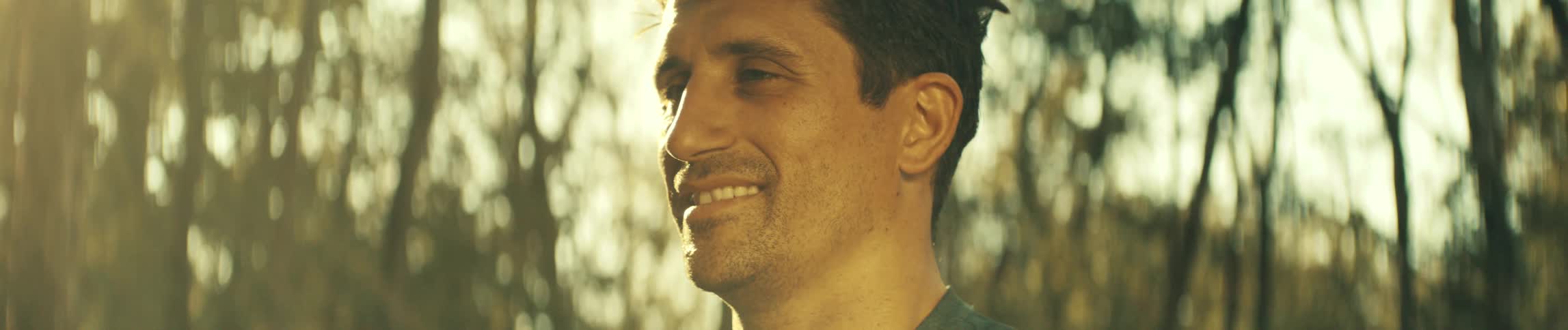 A man with dark hair looks to the side and smiles while standing in a forest with bright golden sunlight and trees in the background during a warm afternoon in a close view