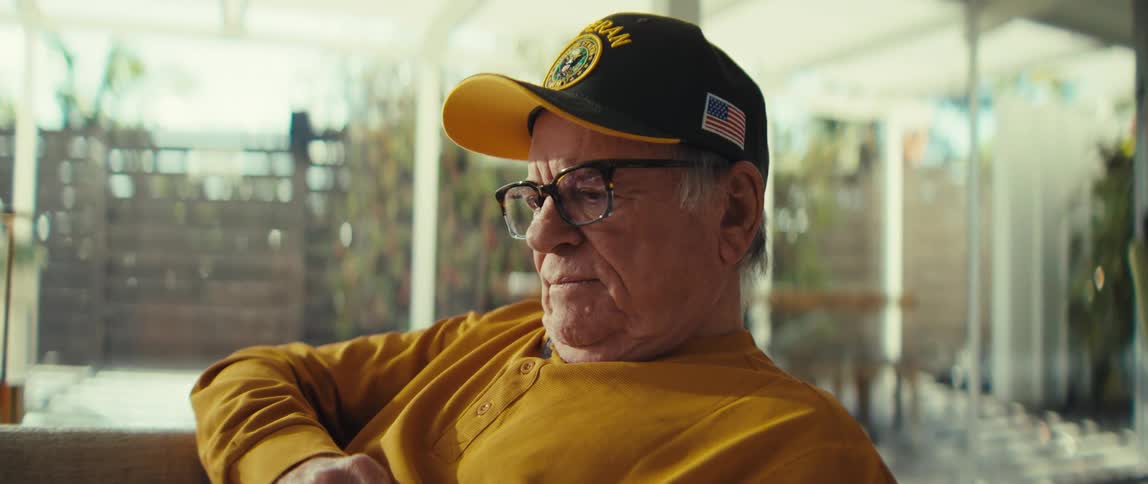 Elderly adult man wearing a yellow shirt and a baseball cap with glasses sitting down on a couch reading next to a window indoors during day time