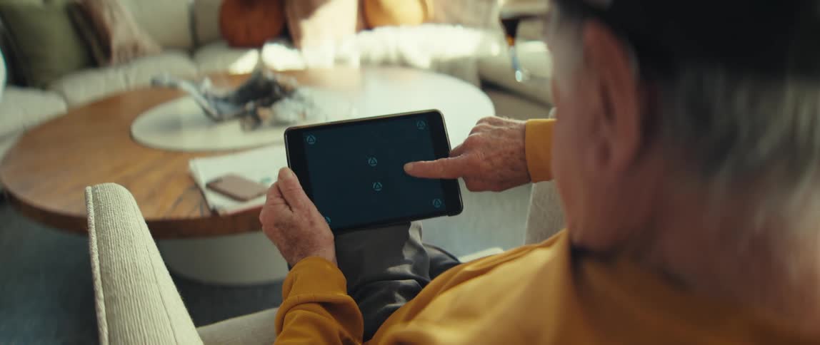 An elderly man sitting behind a coffee table in his living room wearing glasses and scrolling through his phone at midday