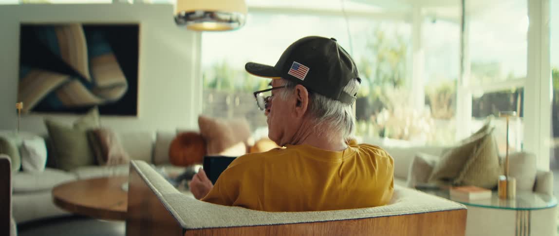An older man wearing a black baseball cap with an american flag patch sits in a chair while looking at a tablet he appears to be deep in thought as he briefly looks away from the screen and touches a pen to his face