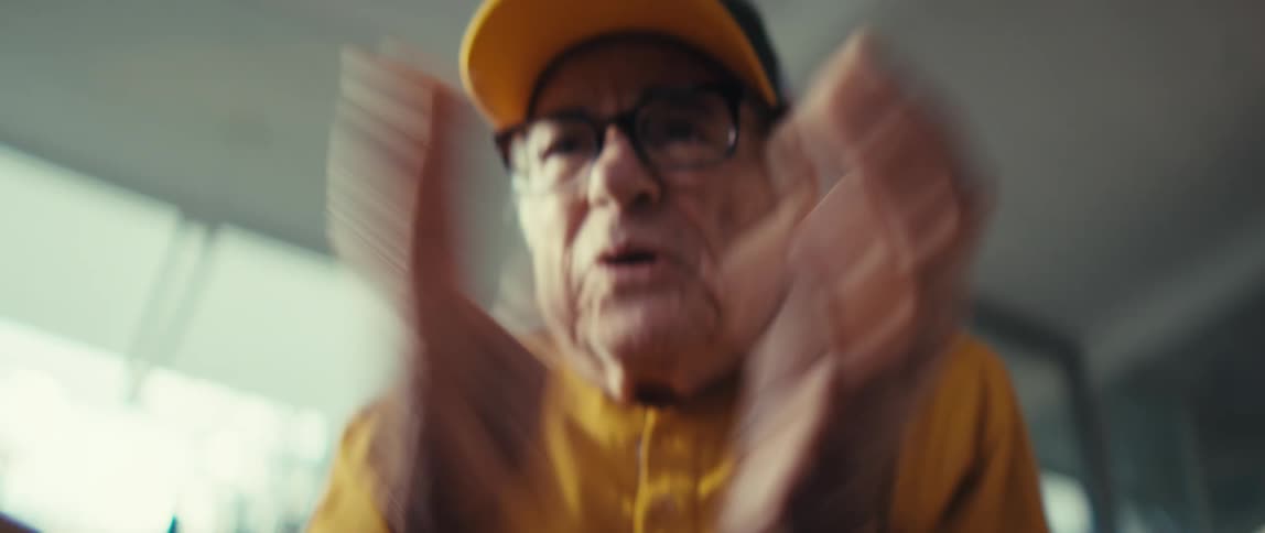 An elderly man wearing a yellow cap and glasses looks directly into the camera while gesturing with his hands during a close up shot inside a brightly lit room with a blurred window background