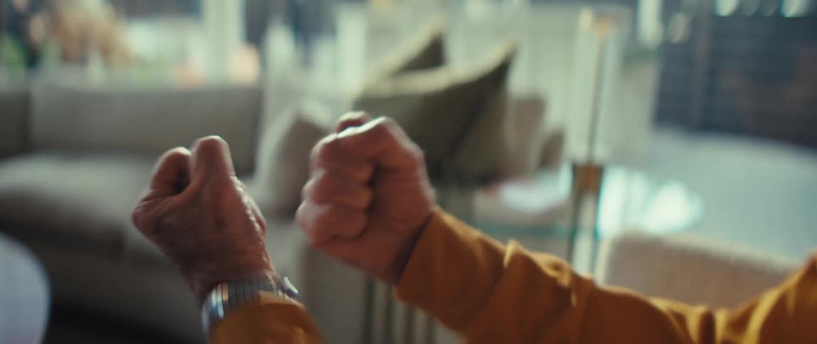An elderly man wearing a yellow sweater sits in a bright living room and raises his clenched fists in a celebratory gesture while a beige sofa and glass table remain blurred in the background