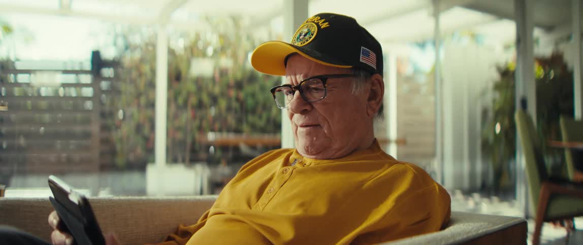 Man wearing a cap and glasses sitting in an armchair while holding a tablet in a room with large windows in the background
