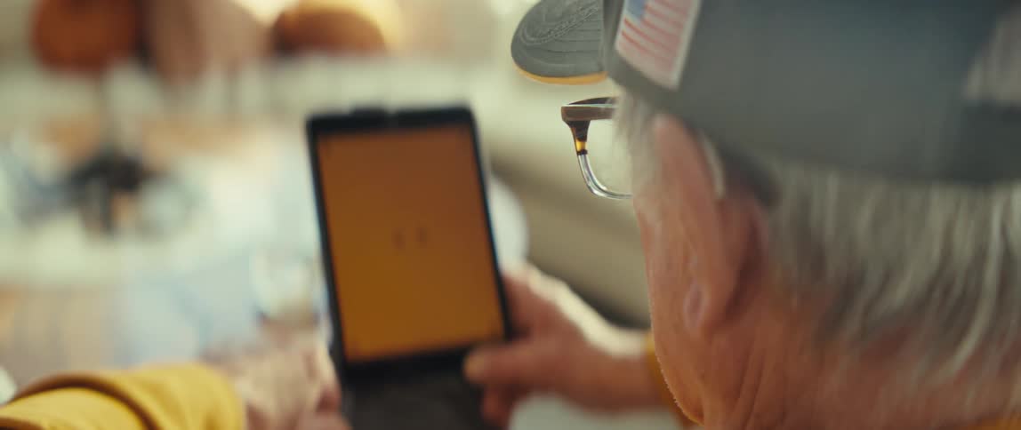 An elderly man wearing a cap and glasses looks intently at a brightly glowing yellow tablet screen gently holding the device in a warmly lit living space
