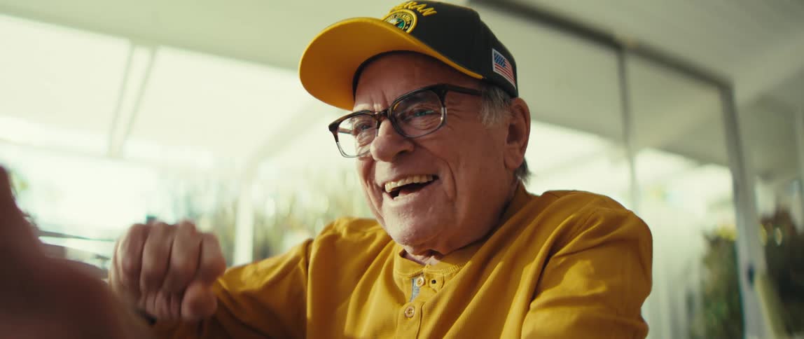 An elderly man sits in a living room cheering and smiling as he looks at a tablet he his holding