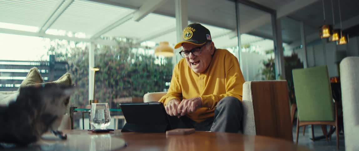 Elderly man wearing a cap sitting in an armchair clapping in front of a tablet on the table with a cat nearby