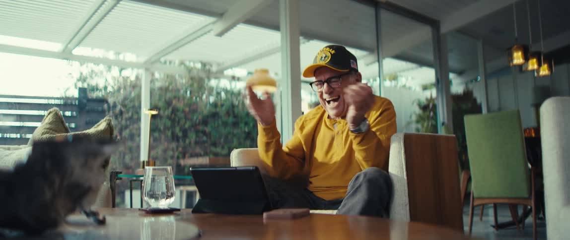 An elderly man wearing a yellow sweater and a veteran hat sits in a modern living room cheering and pumping his fists with excitement while looking at a tablet on a table