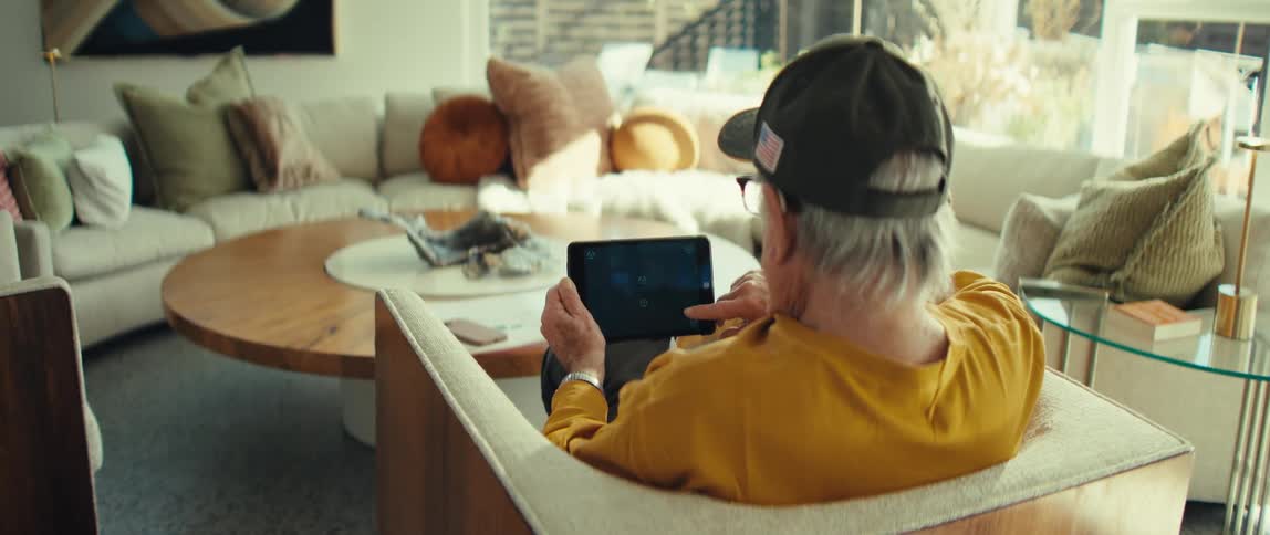 A man sitting on a chair in the living room with a coffee table in front of him wearing a yellow tshirt and a black baseball cap scrolling through his tablet with glasses on at midday