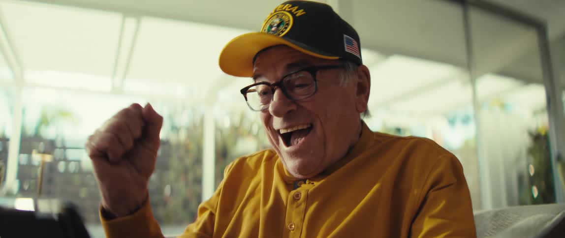 Elderly man wearing a cap and glasses sitting in an armchair reacting with surprise while looking at a tablet