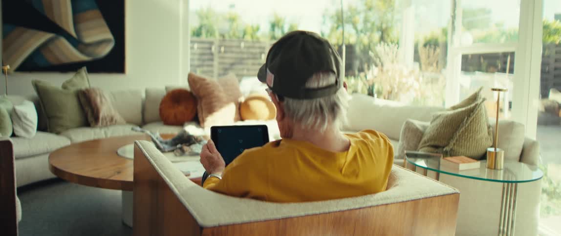 An older man wearing a brown hat and a yellow shirt sits in an armchair while using a tablet in a bright and modern living room in a back view