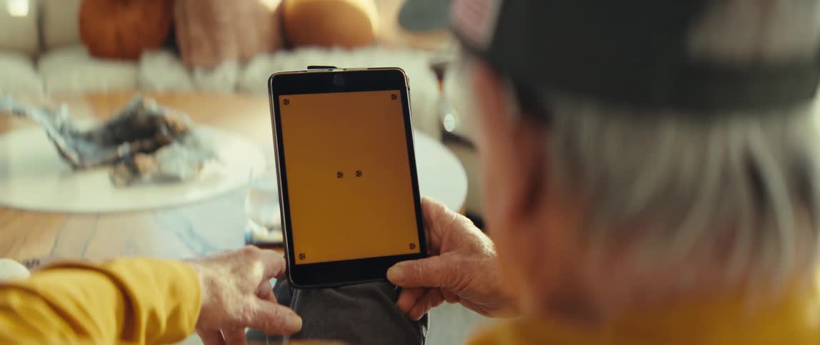 An older man wearing a baseball cap and yellow shirt holds a small tablet with a bright yellow screen in a sunlit room