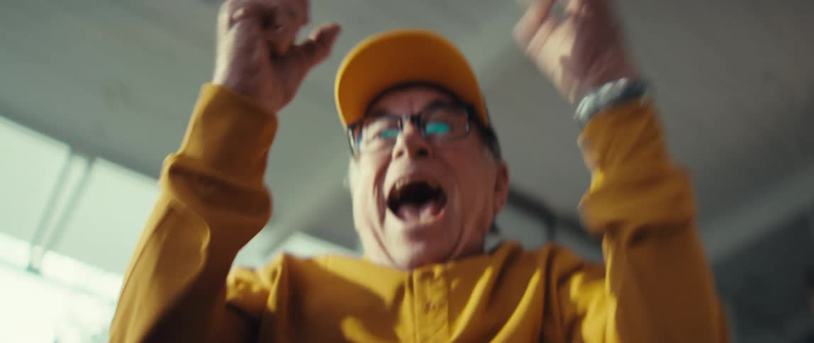 An elderly man in a yellow cap and shirt watches something intently with a look of mounting anticipationhe suddenly erupts into a joyful celebration shouting and raising his fists in victory with a wide smile